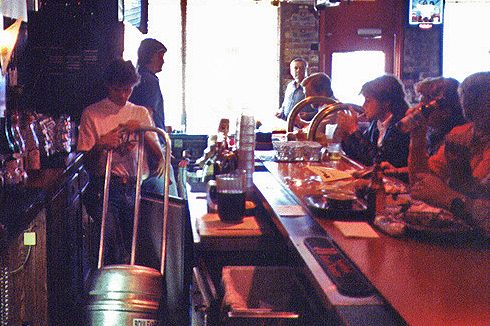 Boulevard Brewing Celebrating 35 Years 26 John McDonald in a white shirt moves a keg behind the bar, while several patrons sit at the counter drinking and conversing about their day. A person stands silhouetted in the entrance, and the interior is dimly lit, creating a cozy atmosphere.