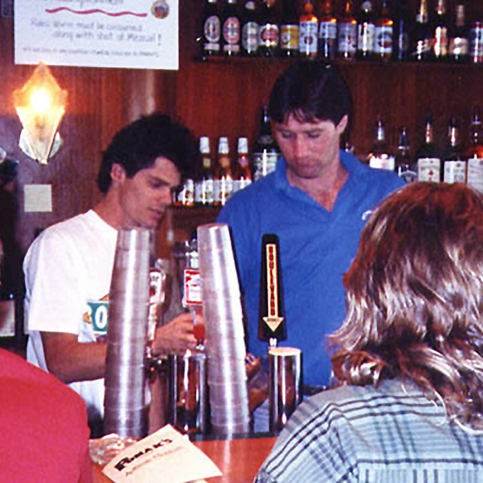 Boulevard Brewing Celebrating 35 Years 27 Two men are working behind a bar. The man on the left is stacking clear plastic cups while the man on the right, in a blue shirt, is holding a drink mixer. Various bottles are displayed on the shelf behind them. In the foreground, about to serve customers, stands a person with long hair and a pamphlet.