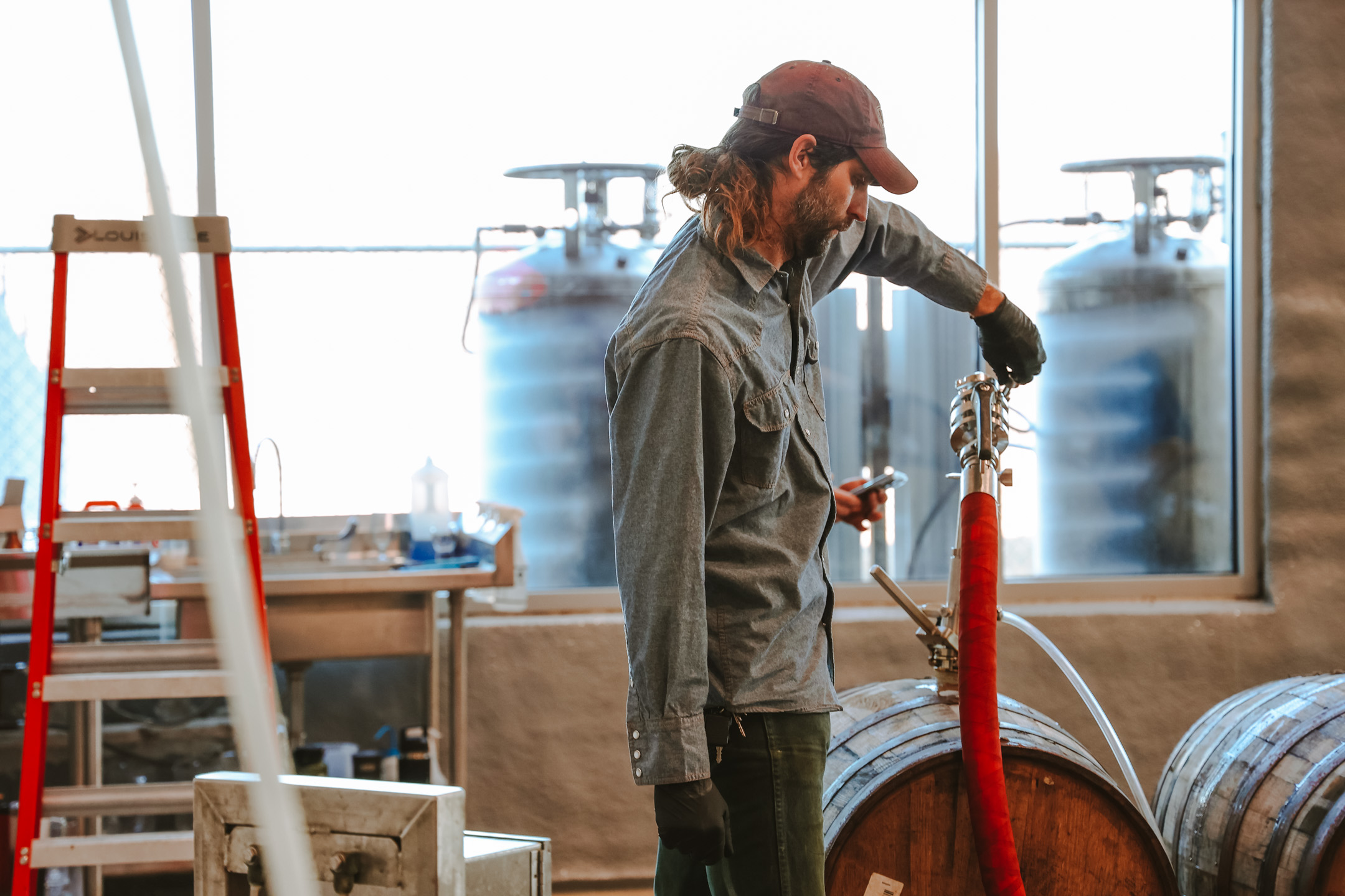 Boulevard Introduces Three Jokers – Naturally Soured Imperial Wit Ale Aged in Chardonnay Barrels 8 A person wearing a brown cap and blue work shirt operates a brewery hose connected to a wooden barrel. Behind them, there are large stainless steel tanks, a ladder, and various equipment, indicating the setting is a brewery or distillery.