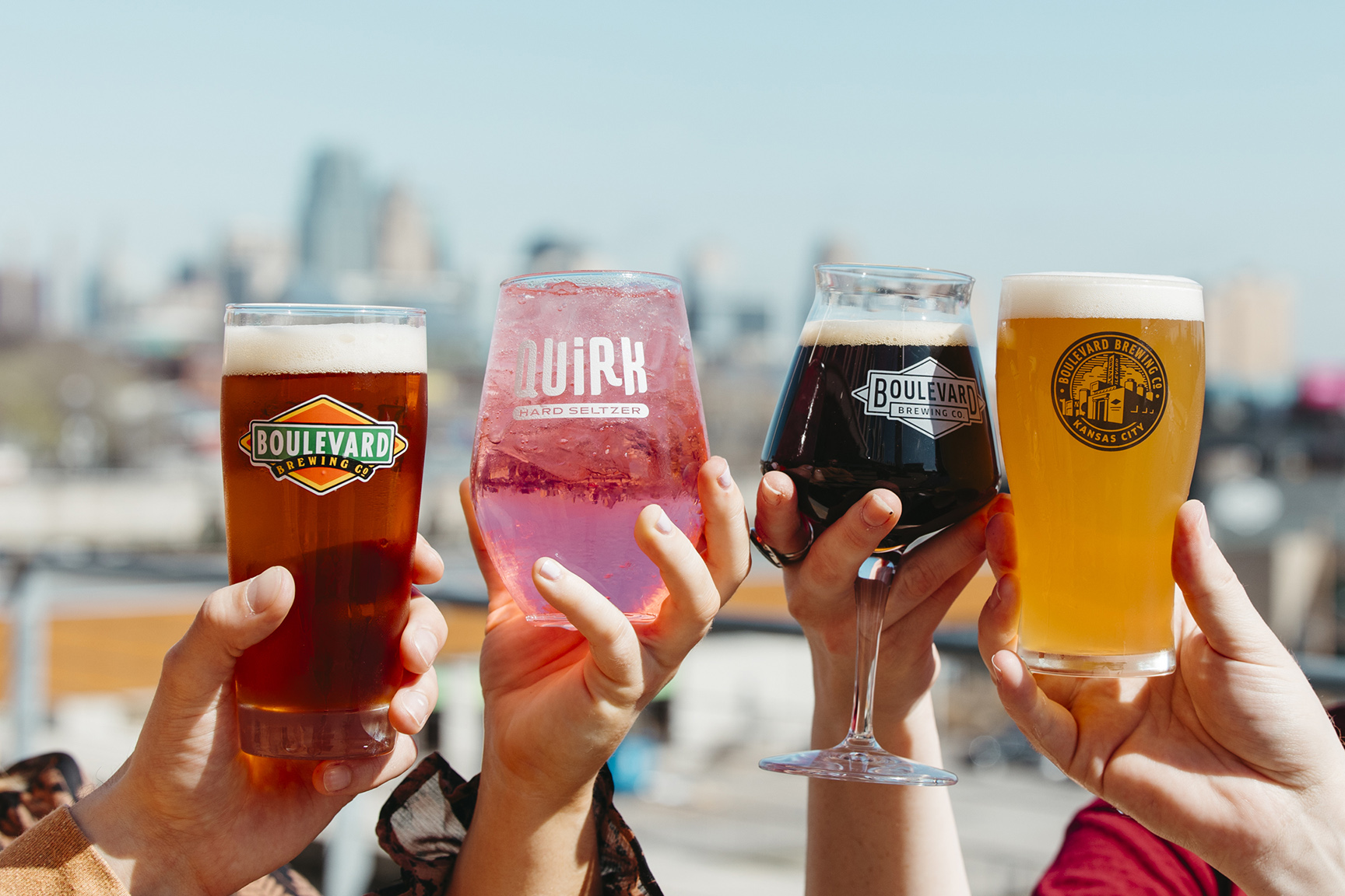 Boulevard Brewing Celebrating 35 Years 36 Four hands holding different types of beverages in branded glasses towards the camera, with a city skyline blurred in the background. The drinks include Pale Ale, Quirk hard seltzer, Bourbon Barrel Quad, and Unfiltered Wheat.