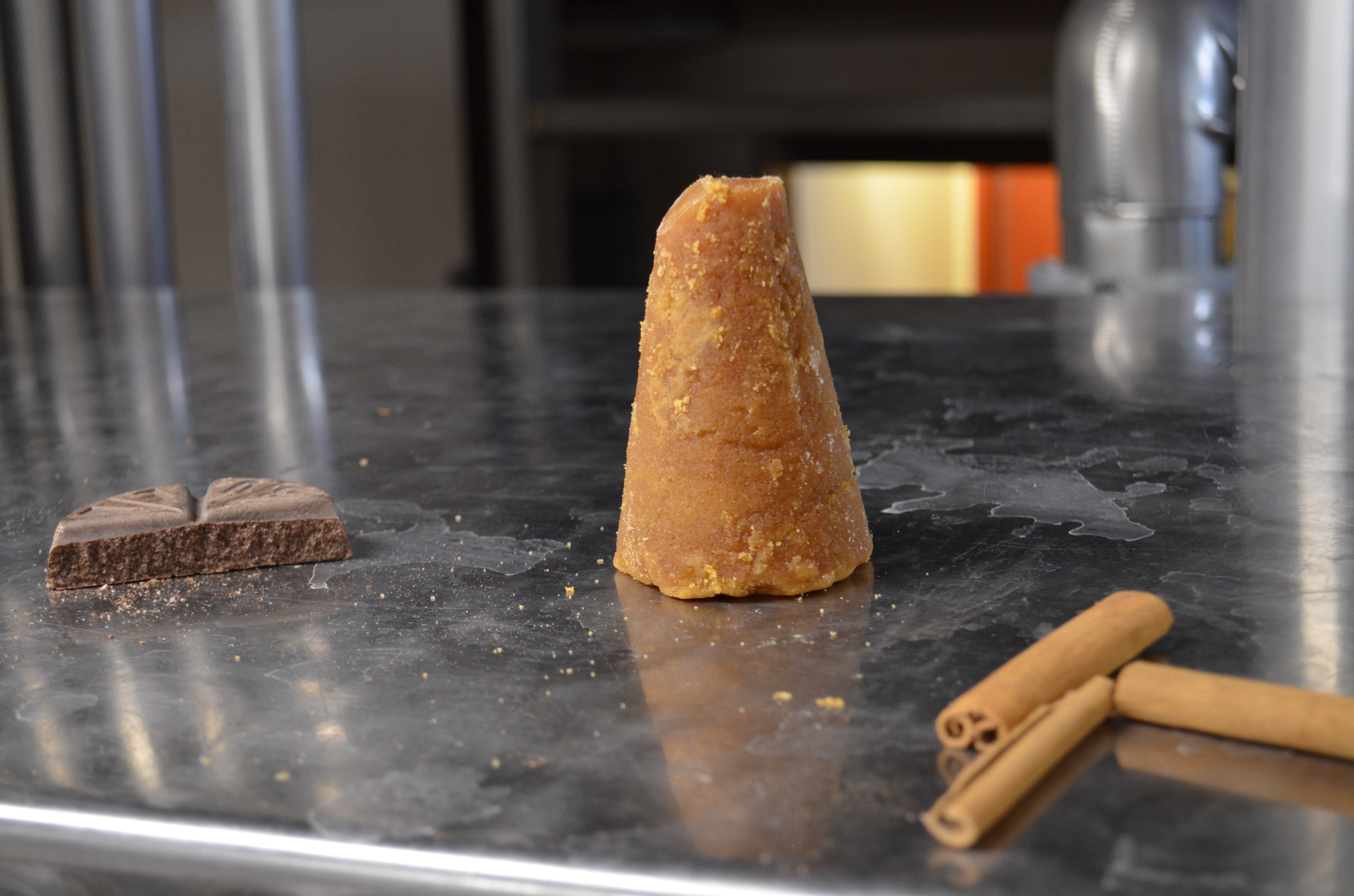 Boulevard Releasing Mexican Xocolatl Ale 19 A cone-shaped piece of brown sugar (piloncillo) stands on a shiny metal surface, surrounded by a bar of chocolate and several cinnamon sticks. The background is slightly blurred, suggesting an indoor setting.