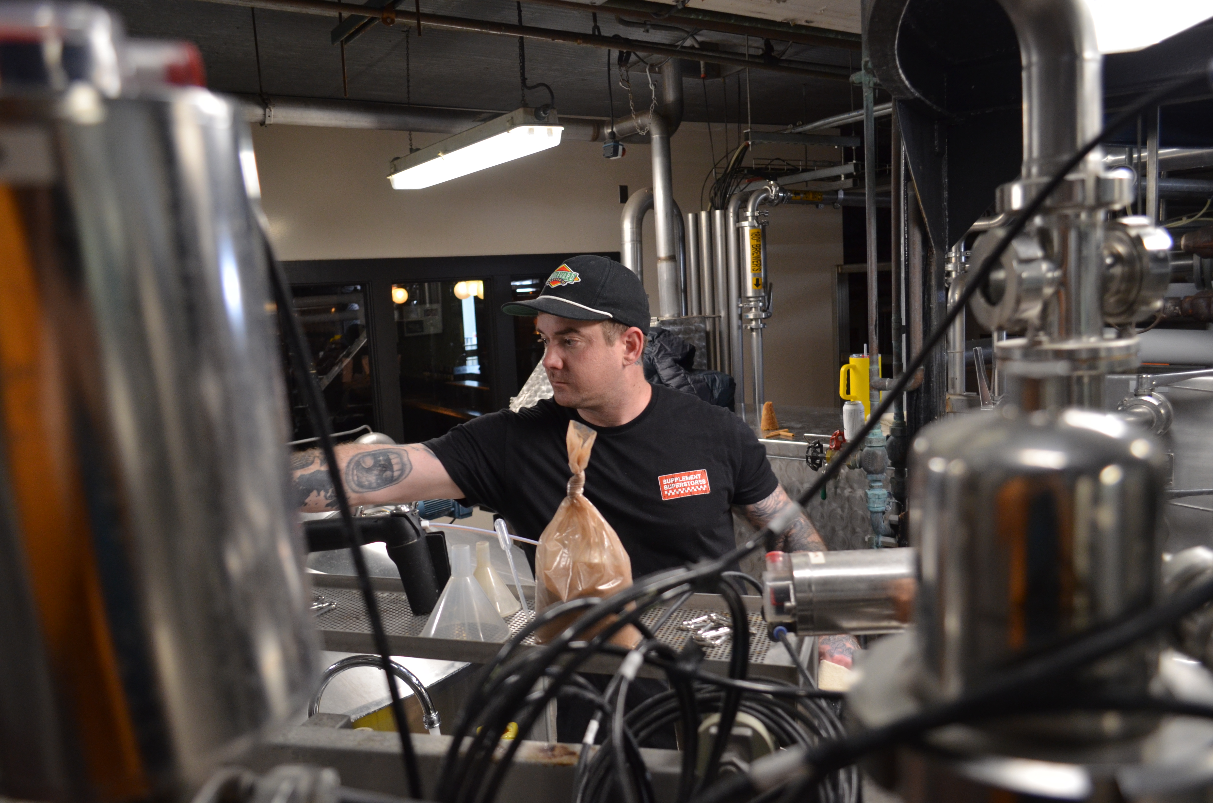 Boulevard Releasing Mexican Xocolatl Ale 24 A person in a black cap and t-shirt works amidst industrial brewing equipment. They are adjusting machinery, surrounded by metal pipes and containers. A beige sack and coiled wires are visible in the foreground.
