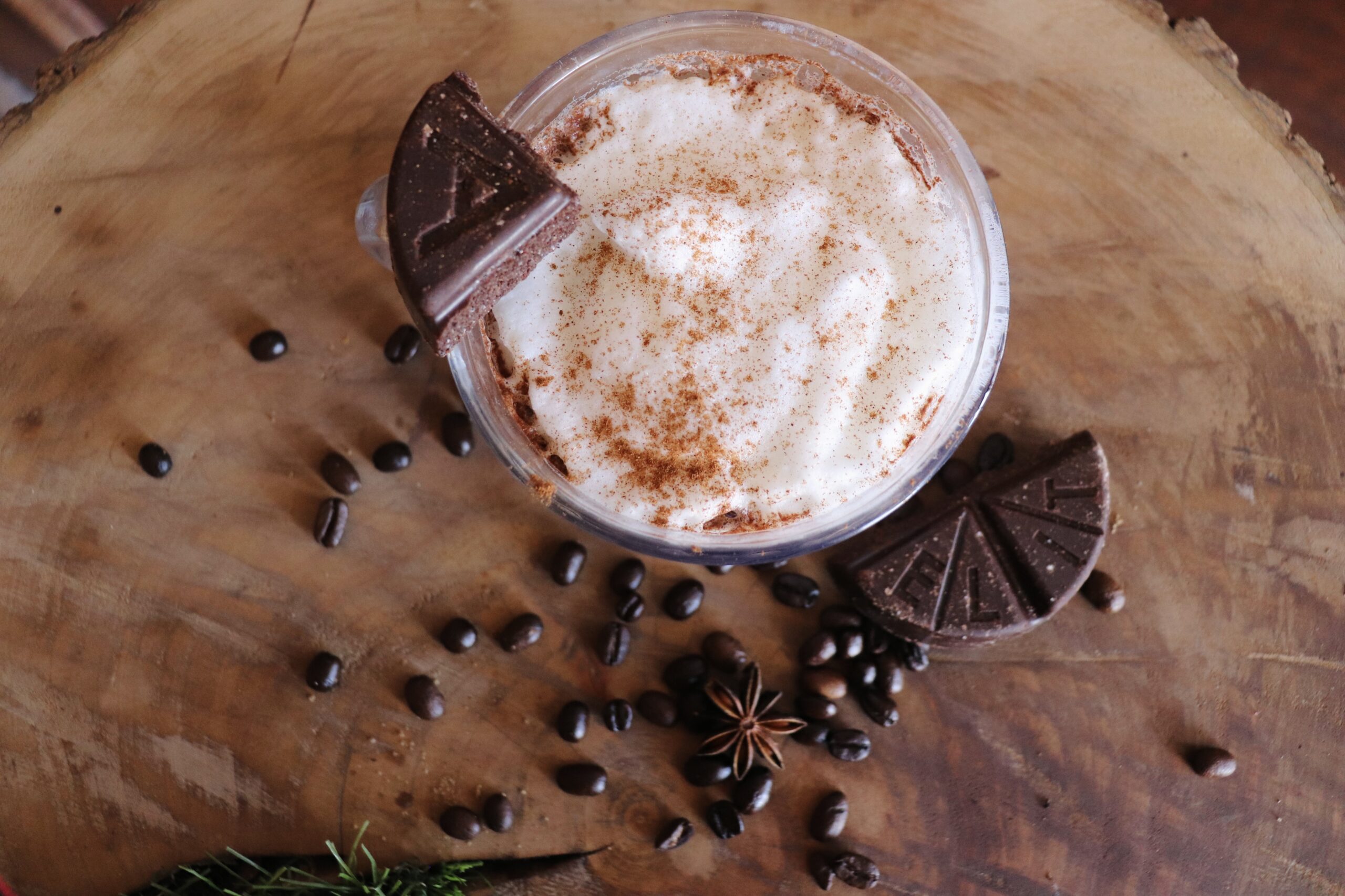 Boulevard Releasing Mexican Xocolatl Ale 18 A creamy coffee drink topped with whipped cream and cinnamon, served in a glass mug. Garnished with triangular pieces of dark chocolate and surrounded by coffee beans, anise stars, and greenery on a wooden surface.