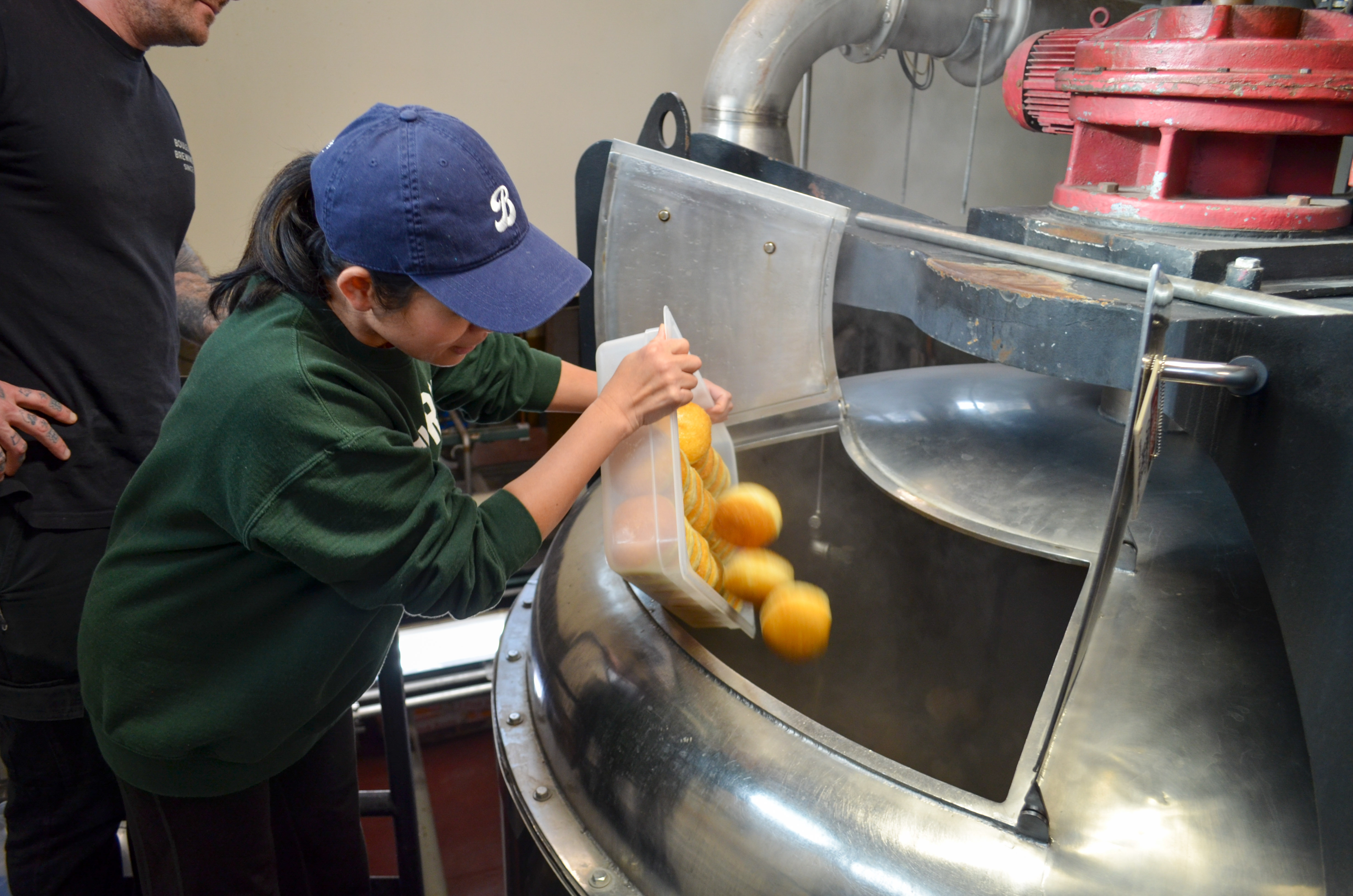 Boulevard Releasing Coconut Pandan Doughnut Ale Collaboration 12 A person wearing a blue cap and green sweater is adding oranges into a large industrial machine, observed by another person. The scene appears to be in a factory setting with stainless steel equipment.