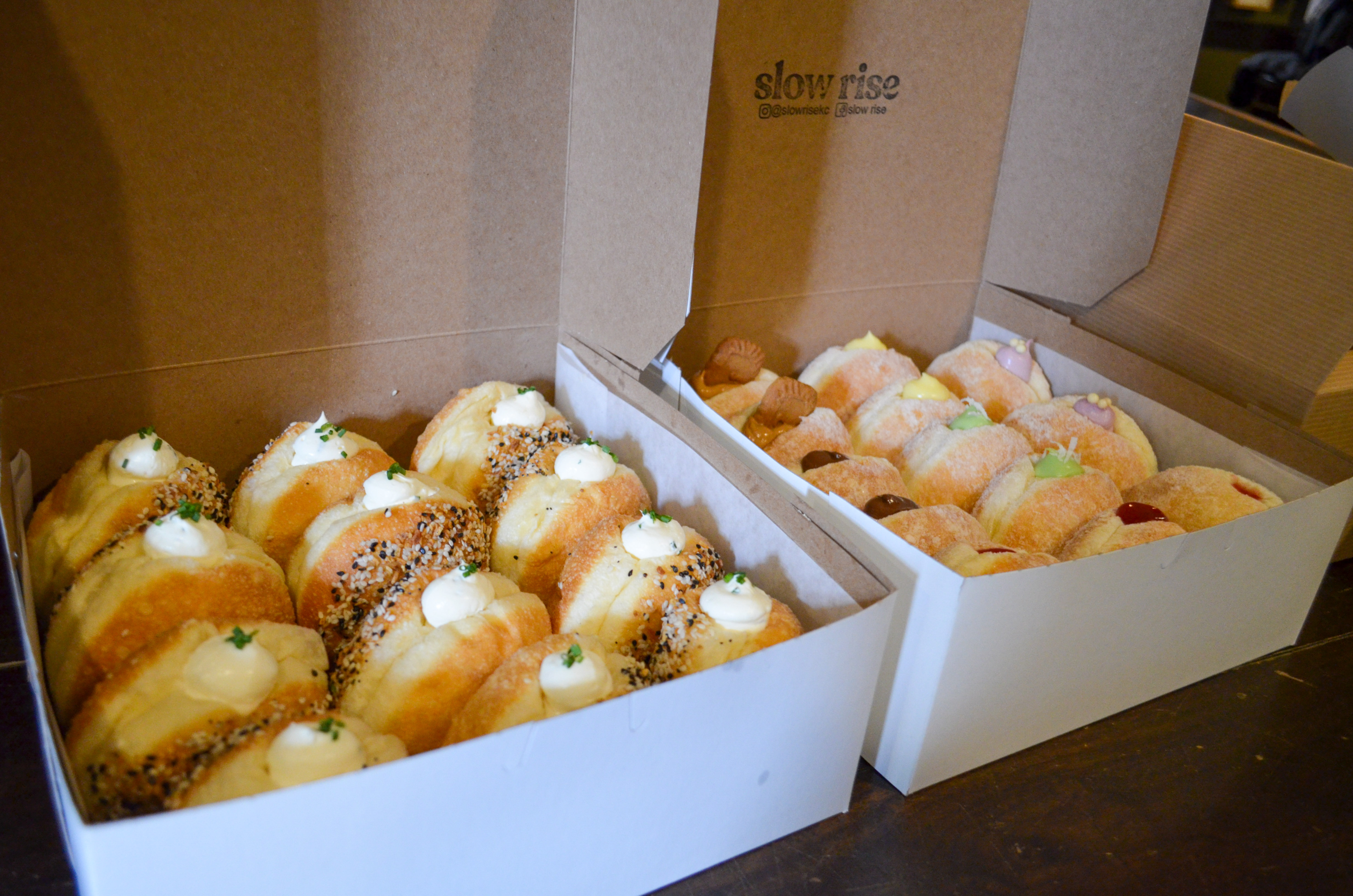 Boulevard Releasing Coconut Pandan Doughnut Ale Collaboration 11 Two open boxes filled with assorted pastries on a wooden table. The left box has pastries topped with cream and herbs, sprinkled with seeds. The right box contains smaller pastries, some topped with powdered sugar or jam.