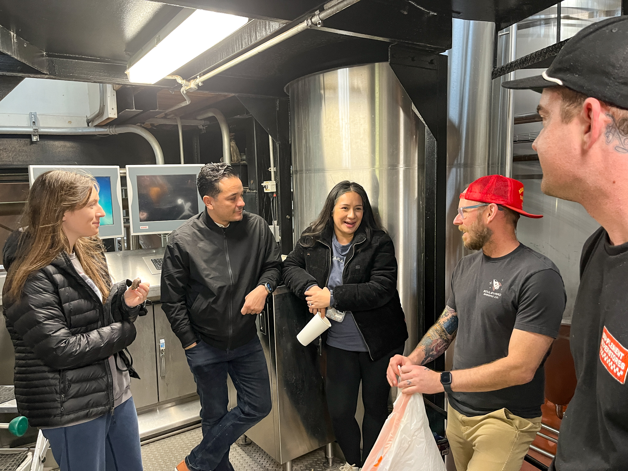 Boulevard Releasing Mexican Xocolatl Ale 25 A group of five people are gathered in an industrial setting with metal equipment and monitors in the background. Two of them, a man and a woman, are engaged in conversation while others listen attentively. They are dressed casually and appear relaxed.