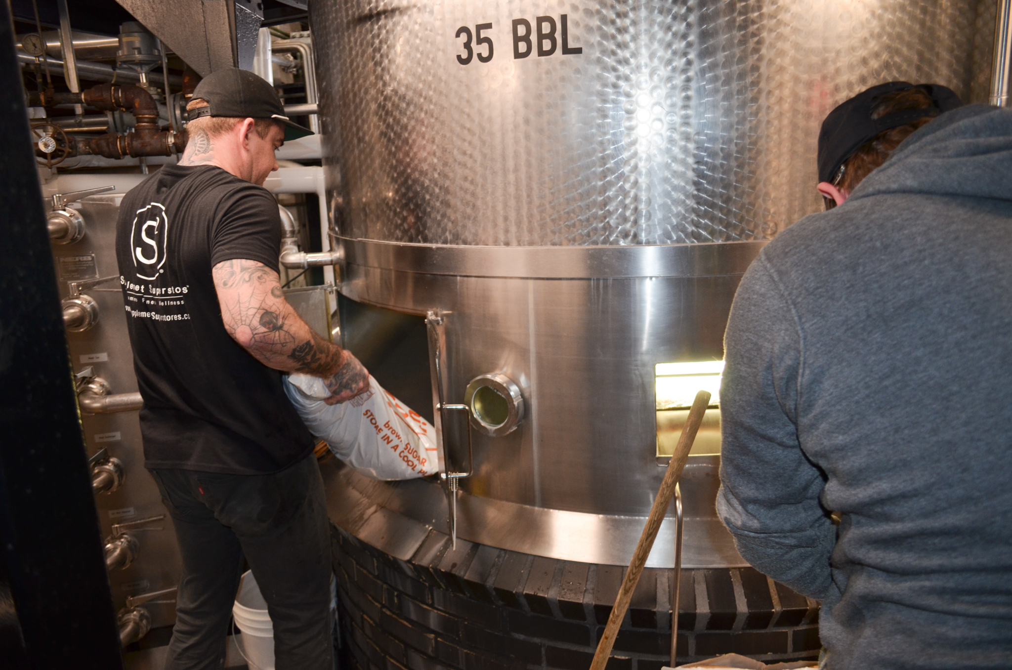 Boulevard Releasing Mexican Xocolatl Ale 22 Two people work near a large stainless steel brewing tank labeled "35 BBL." One person is pouring ingredients, and the other is tending to the tank. Pipes and equipment surround them in the brewery.