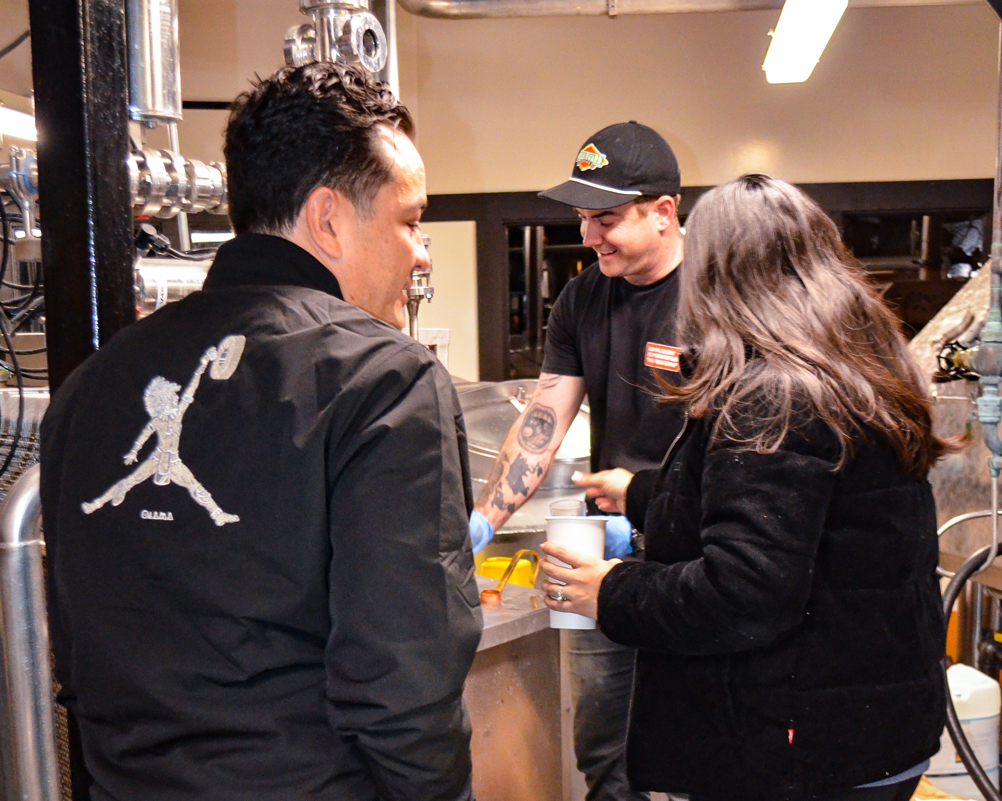 Boulevard Releasing Mexican Xocolatl Ale 26 Three people are gathered in a workshop setting. One man, wearing a black cap and displaying tattoos, is engaging with the others, who are wearing black jackets. They seem to be involved in a collaborative activity.