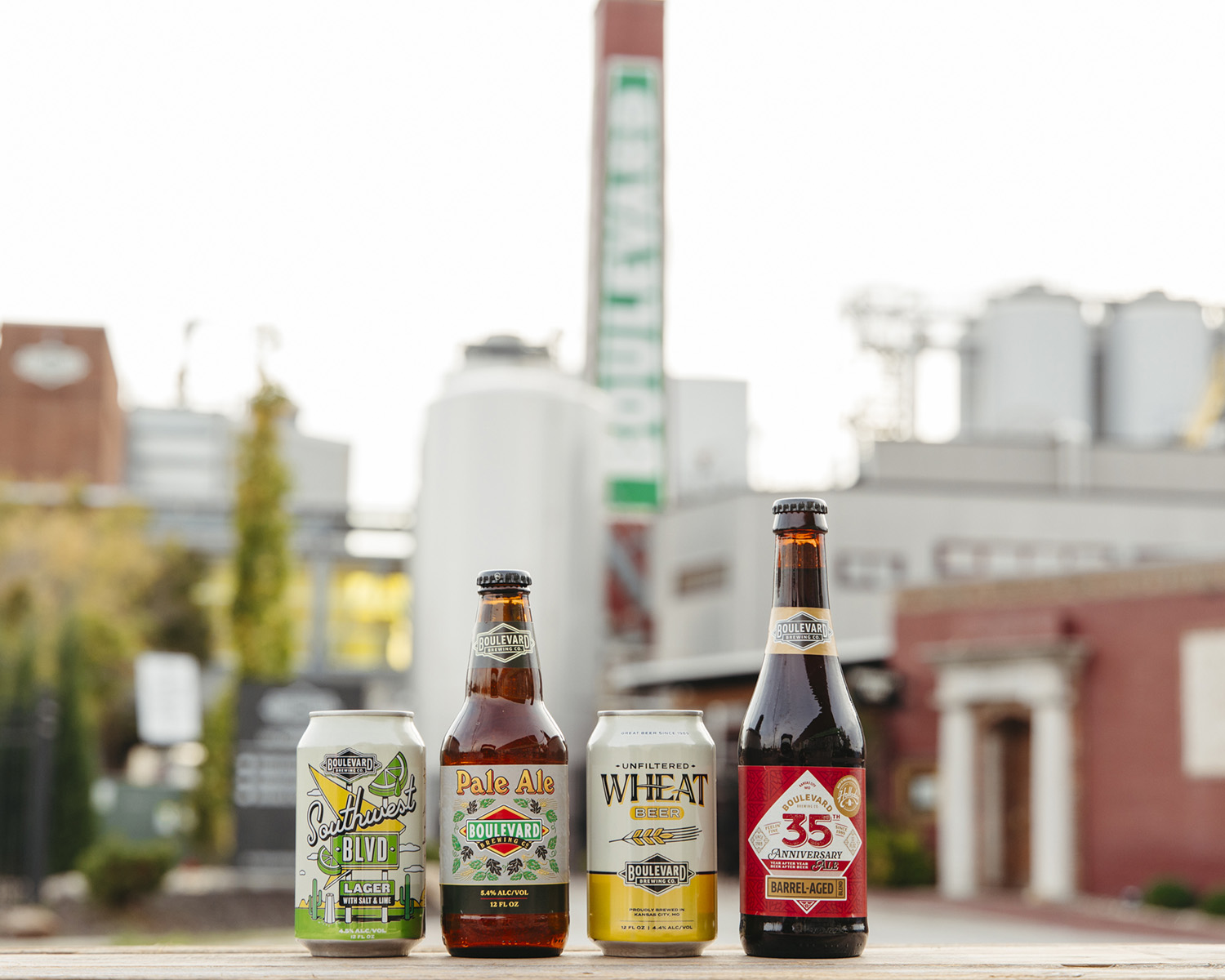 Boulevard Brewing Celebrating 35 Years 35 Four Boulevard Brewing Company beer bottles and cans are displayed on a wooden surface. The backdrop features the brewery's industrial buildings, including a tall chimney with the Boulevard logo. The setting conveys a craft beer atmosphere.