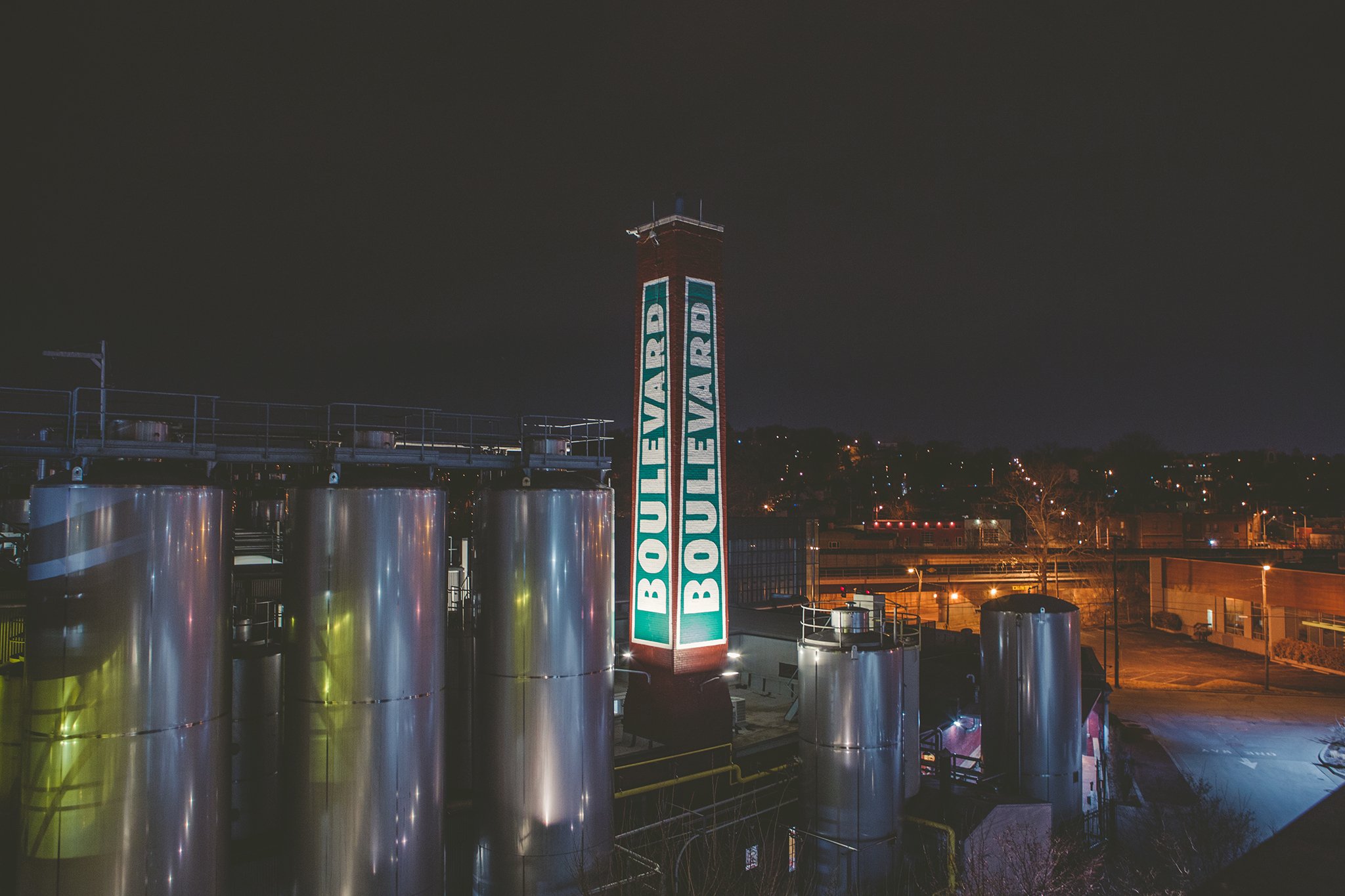 Boulevard Brewing Celebrating 35 Years 25 Night view of an industrial facility with large metal tanks and a tall illuminated chimney displaying the word "Boulevard" repeatedly. City lights are visible in the background.