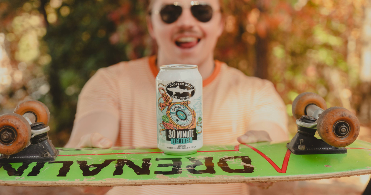 Dogfish Head Adding 30 Minute Light IPA 14 A 12 oz can of Dogfish Head 30 Minute Light IPA balanced on a skateboard with a person posing behind it