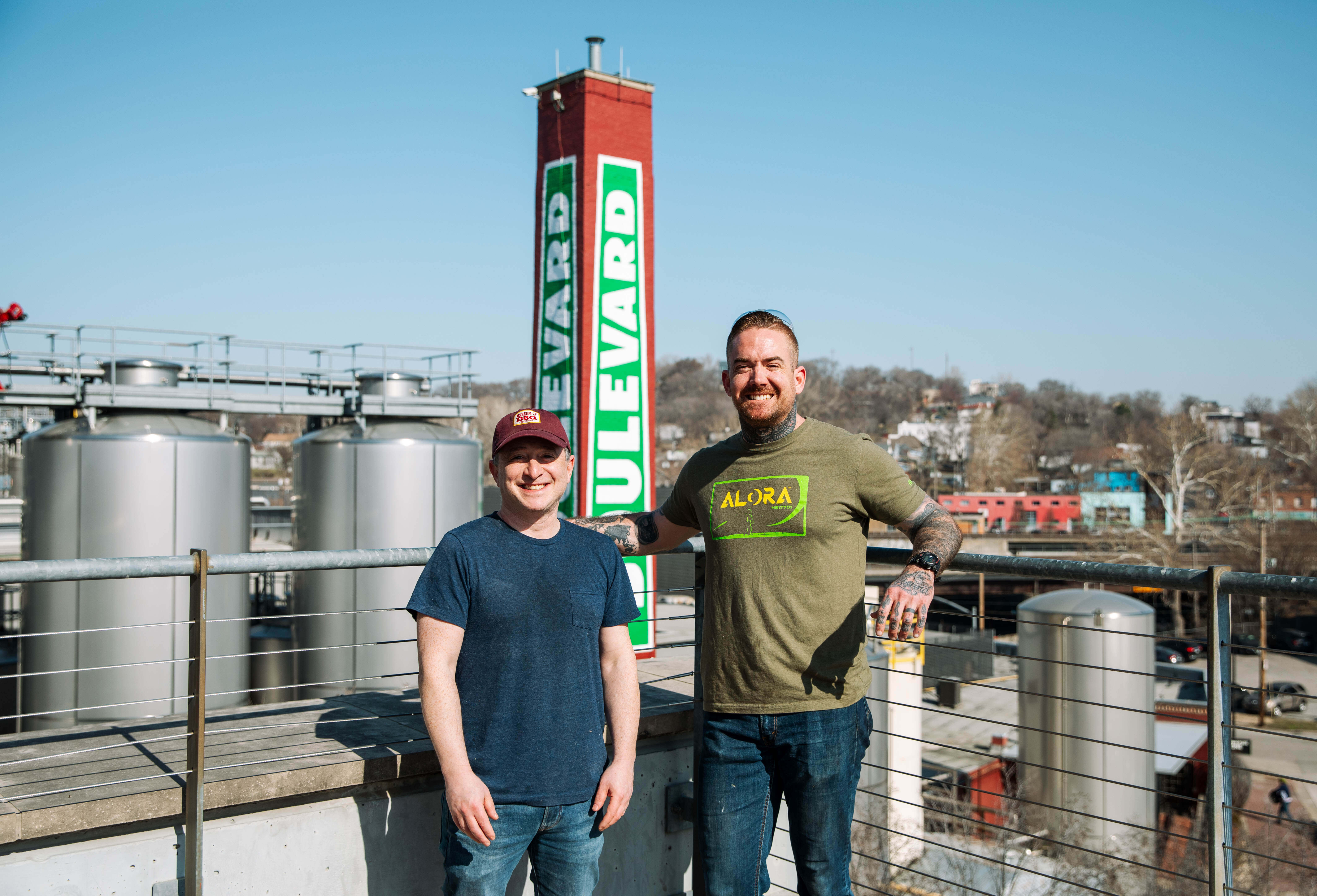Boulevard Brewing Releasing Brewhouse One Series Mo’ BBQ Two men stand smiling on an outdoor terrace with large brewing tanks and a tall red and green "Boulevard" sign behind them on a sunny day. One man holds a drink. Buildings and trees are visible in the background.