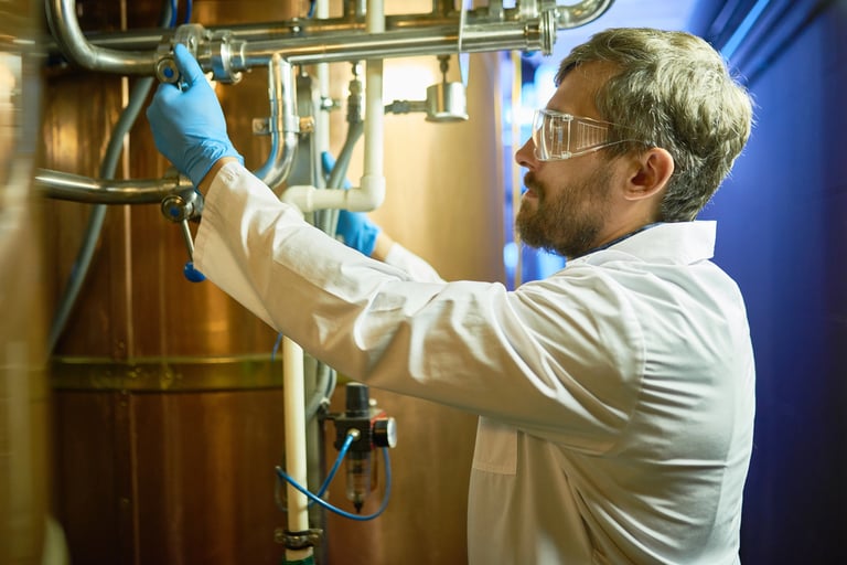 A technician wearing protective goggles and a white coat is adjusting pressure in a cylindrical beer maturation tank while standing at a modern brewing plant.