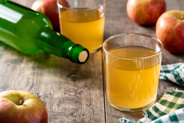 Glass of homemade apple cider with apples and a green bottle on a wooden table, illustrating a beginner’s guide to making cider from apple juice at home.