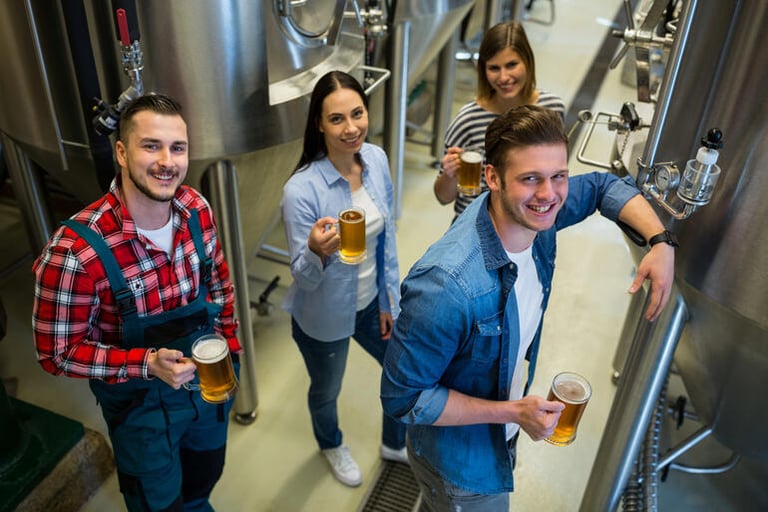 Group of visitors holding craft beer while touring a brewery, capturing memorable moments among stainless steel brewing tanks.