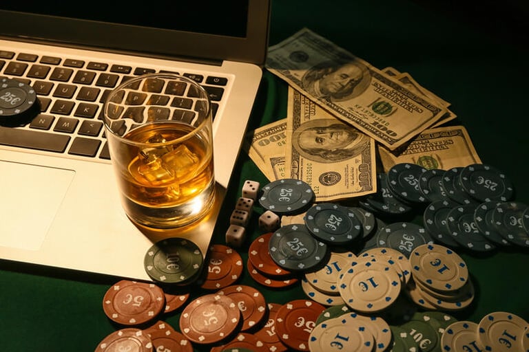 A laptop beside casino chips, cash, dice, and a glass of beer-themed drink.