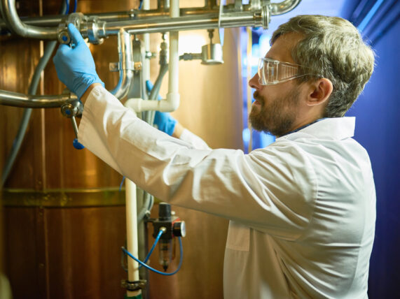 A technician wearing protective goggles and a white coat is adjusting pressure in a cylindrical beer maturation tank while standing at a modern brewing plant.